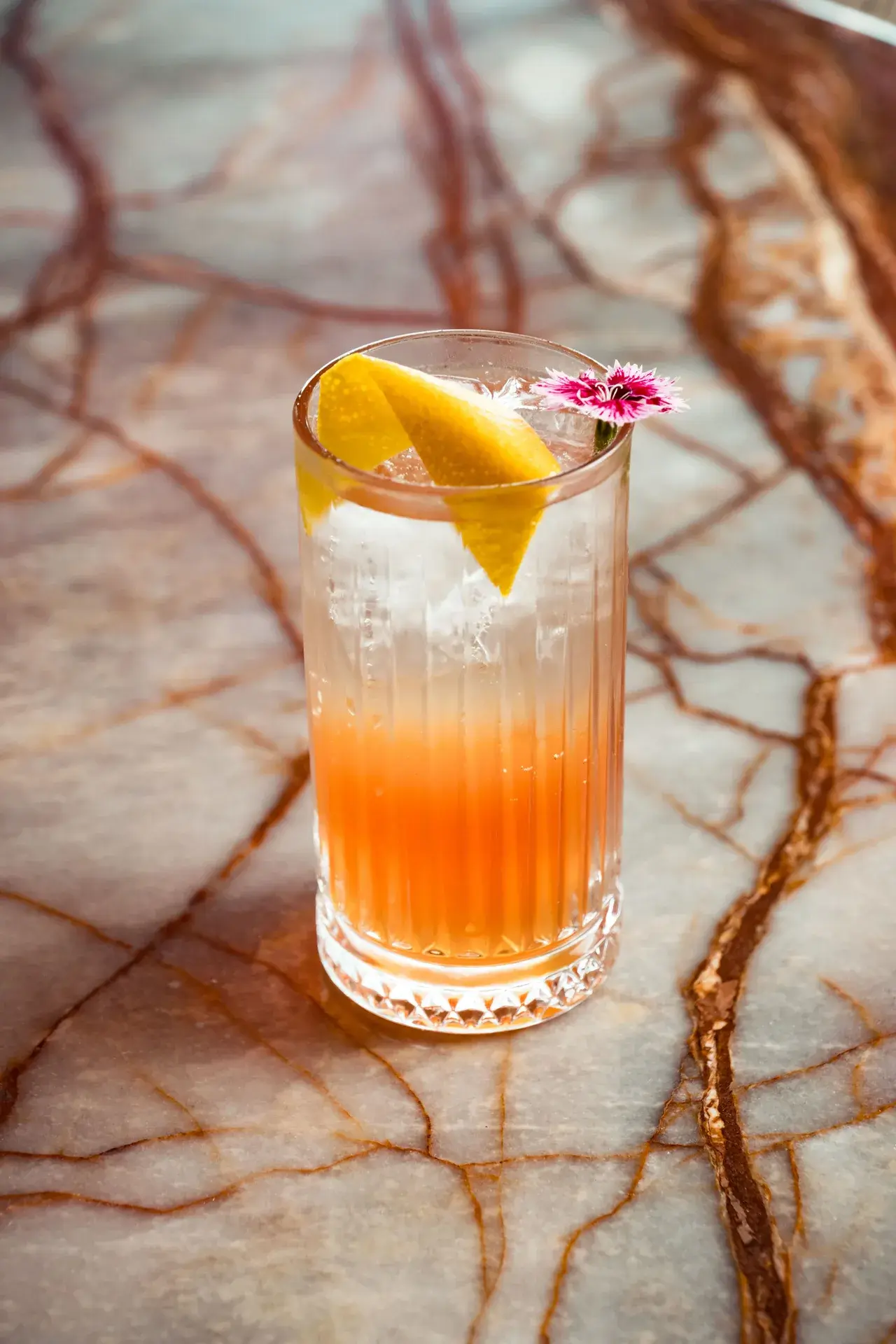 A tall pink citrus cocktail with lemon peel and edible flower garnish on a marbled table
