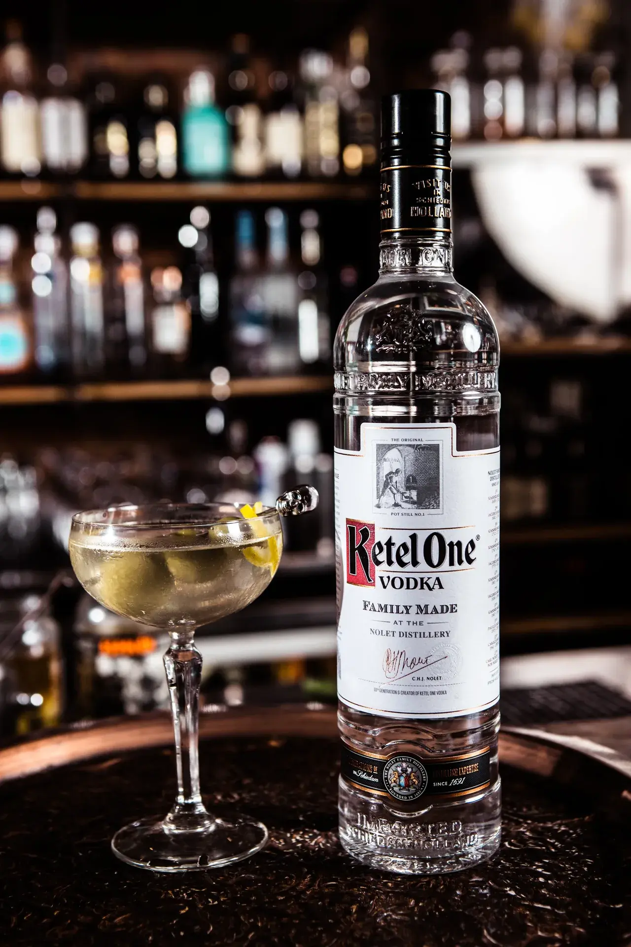 Ketel One Vodka bottle beside a martini garnished with olives on a skull pick in a dimly lit bar