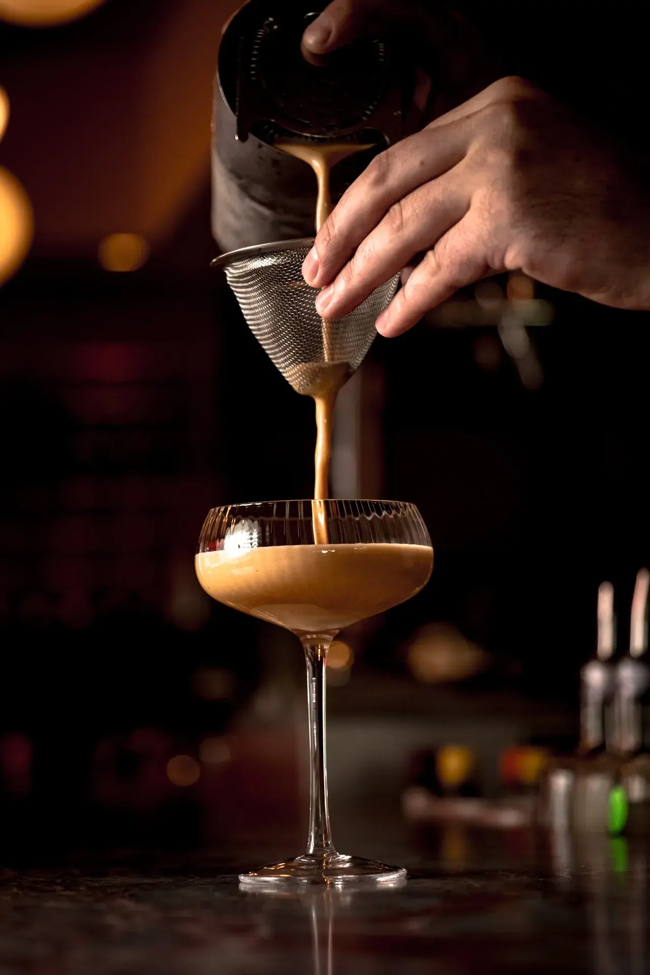 Bartender pouring a creamy espresso cocktail through a fine mesh strainer into a coupe glass