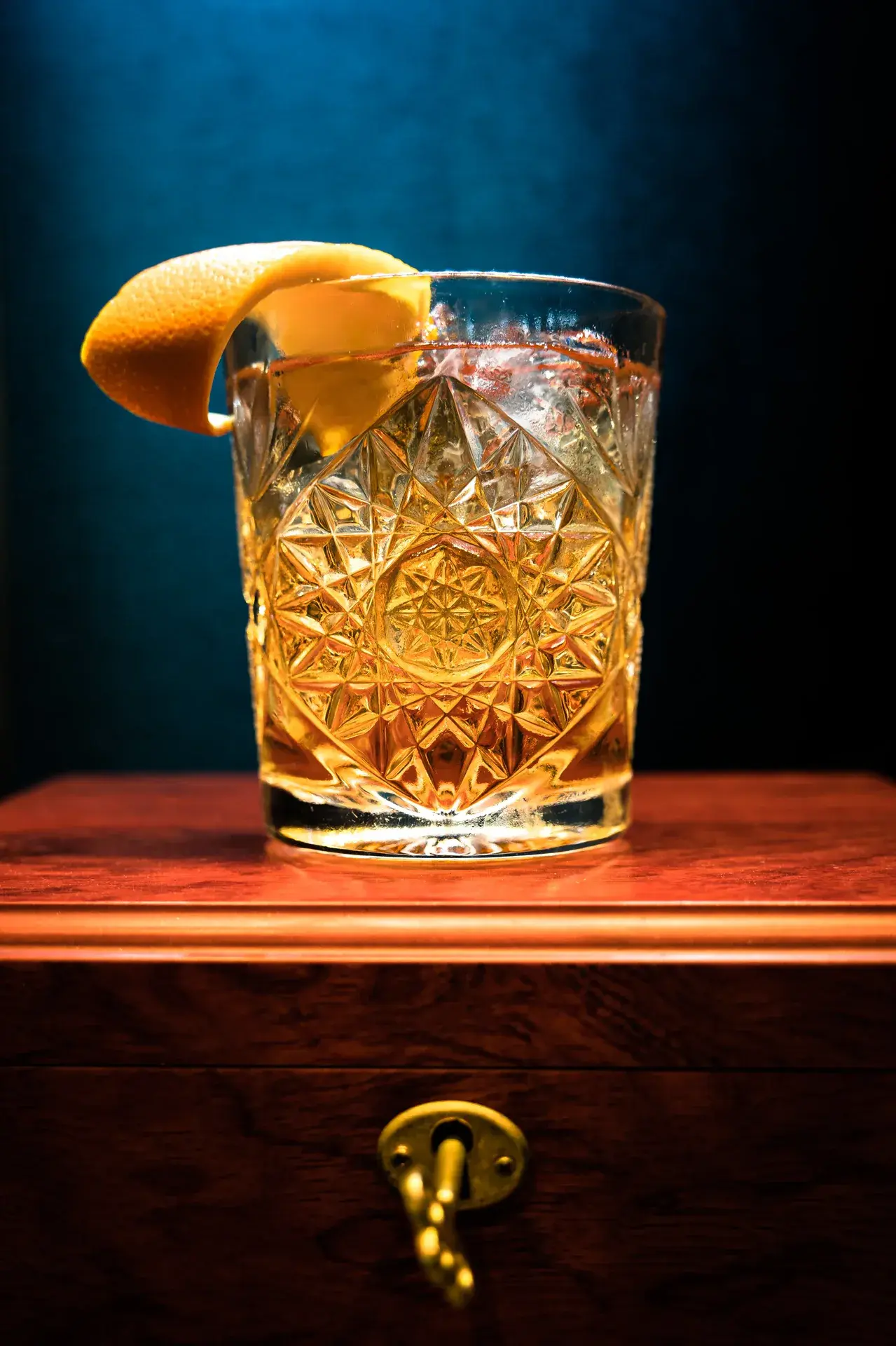 Classic Old Fashioned cocktail in a crystal tumbler with orange peel garnish resting on a wooden cabinet