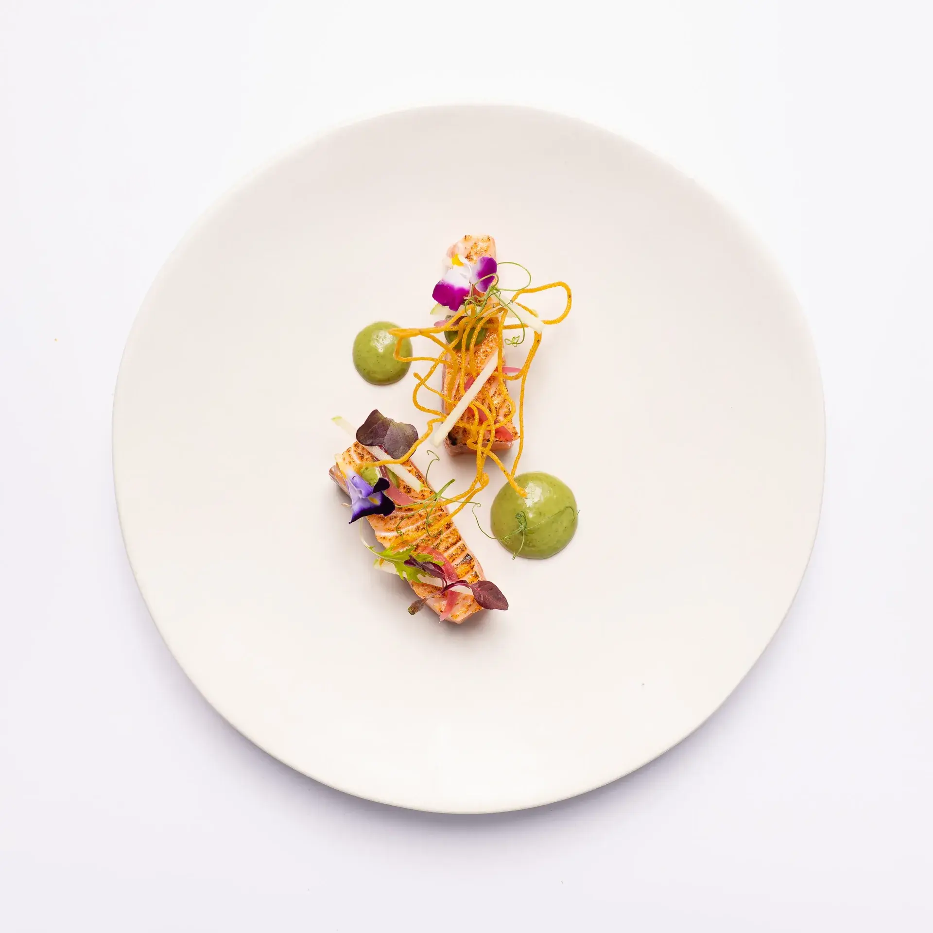 Minimalist plating of seared salmon pieces with edible flowers and vibrant green sauce dots