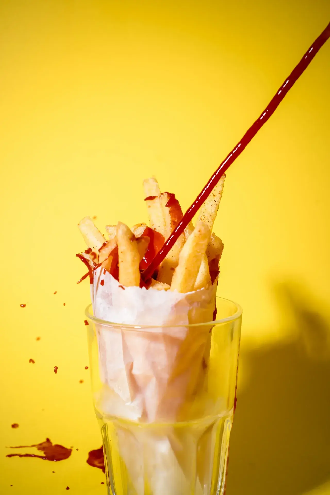 Dynamic high-contrast shot of french fries in a cone with ketchup being squirted mid-air, yellow background