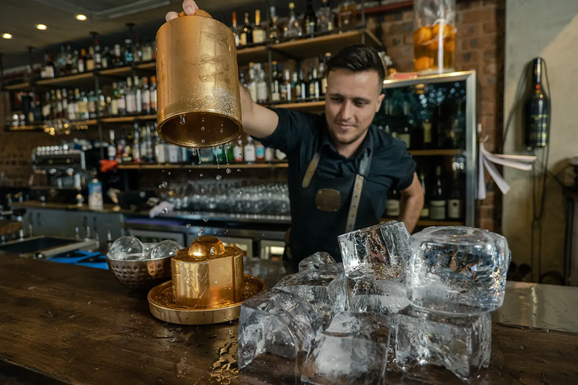 Bartender pouring a drink from a large golden shaker behind clear ice blocks on a bar counter