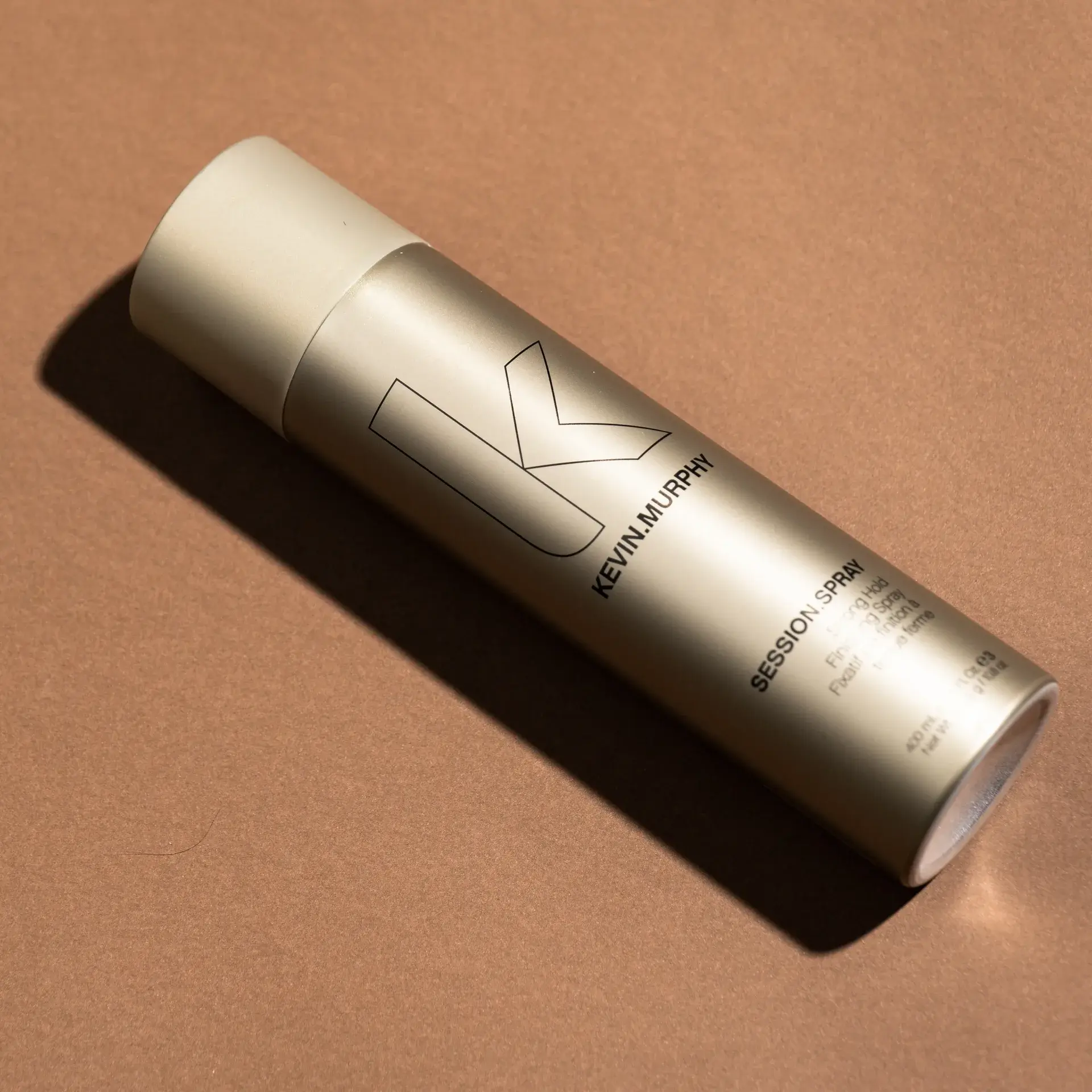 Gold Kevin Murphy Session Spray bottle lying diagonally on a smooth terracotta background